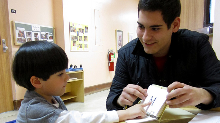 A student pursuing a child development degree sits with a young child who points to the letter ‘M’ on a teaching card.