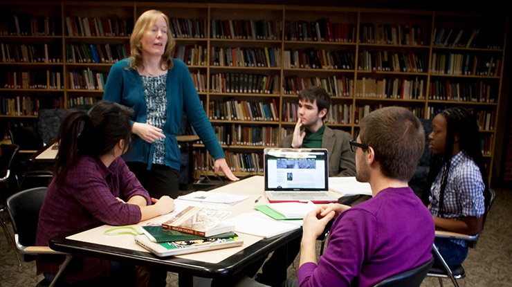 A professor instructs a small table of James Madison College majors in front of a wall of packed bookshelves.