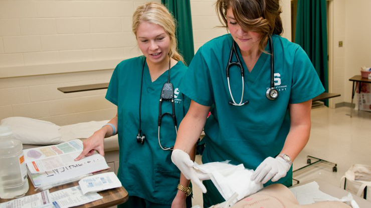 Two students in accelerated nursing programs wear MSU scrubs and consult a textbook for help while using medical supplies. 