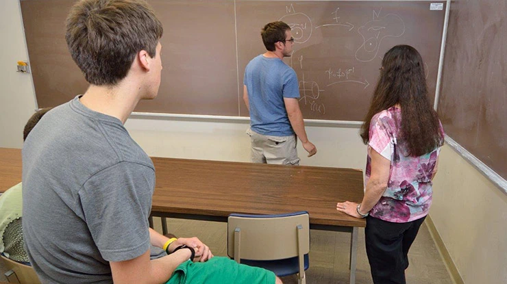 Four math majors face the front of a classroom and examine an advanced mathematics equation written on the chalkboard.