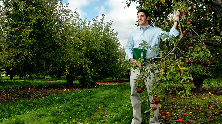 An agribusiness student smiles in an orchard, holding an apple tree branch in one hand and an MSU folder in the other.