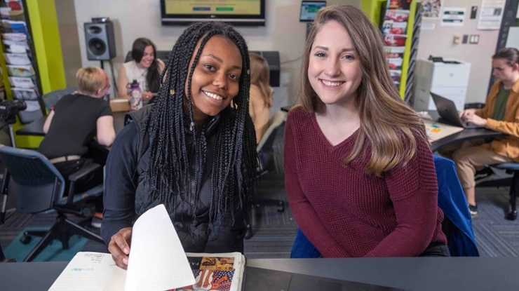Two arts and humanities majors smile as they page through an art book in a student resource center bustling with students.