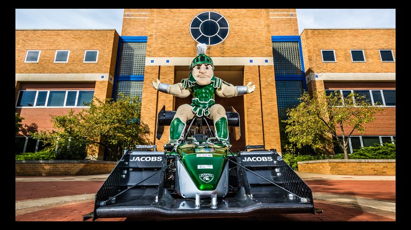 Sparty poses on top of a formula one race car in front of the engineering building.