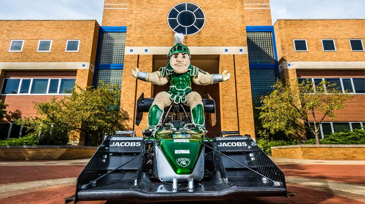 Sparty poses on top of a formula one race car in front of the engineering building.