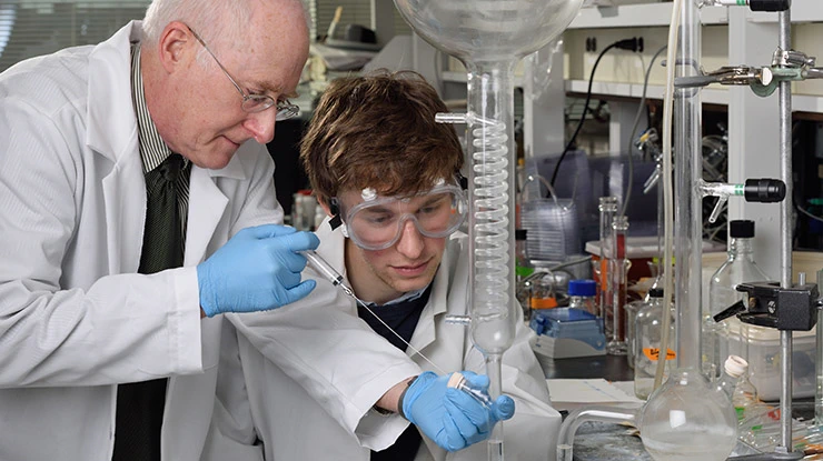 A professor shows a biochemistry major wearing goggles how to use laboratory equipment.