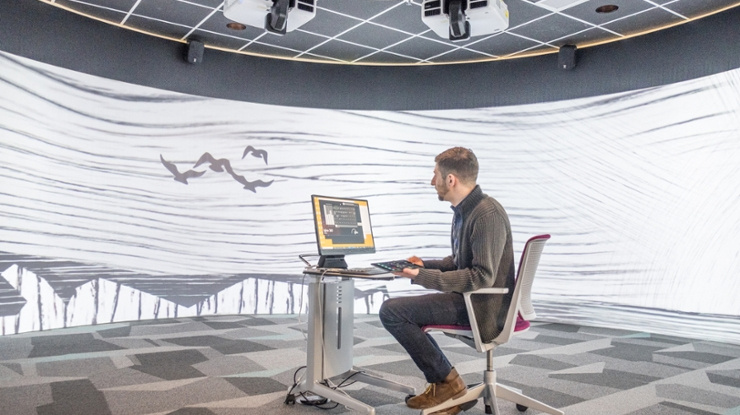 A computational data science student sits at a lone computer station surrounded by a 360° visualization space.
