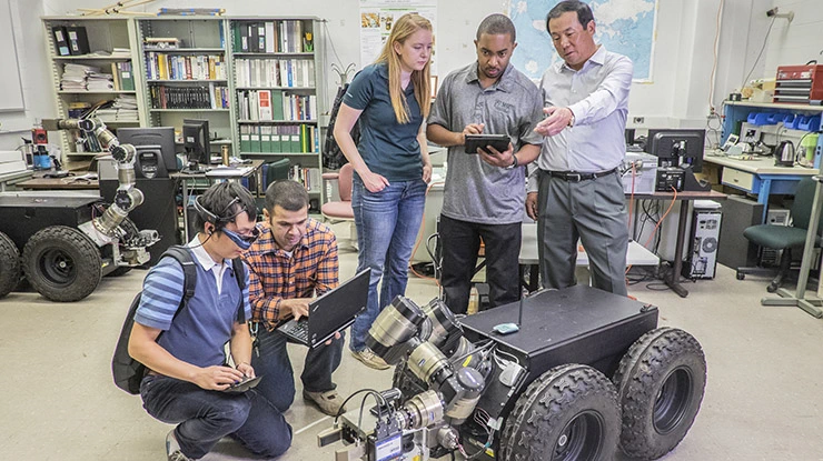 A professor instructs four students pursuing computer engineering degrees on the function of a machine in front of them.