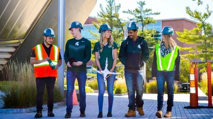 A group of students working on a construction management degree walk next to each other on a sunny day wearing hard hats.