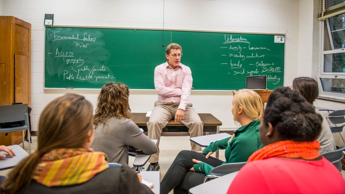 An economic geography professor sits on a table and gives a lecture to a classroom full of economics students.