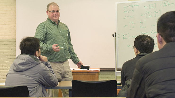 A professor stands next to a whiteboard at the front of a classroom of economics majors, holding a marker and an eraser.