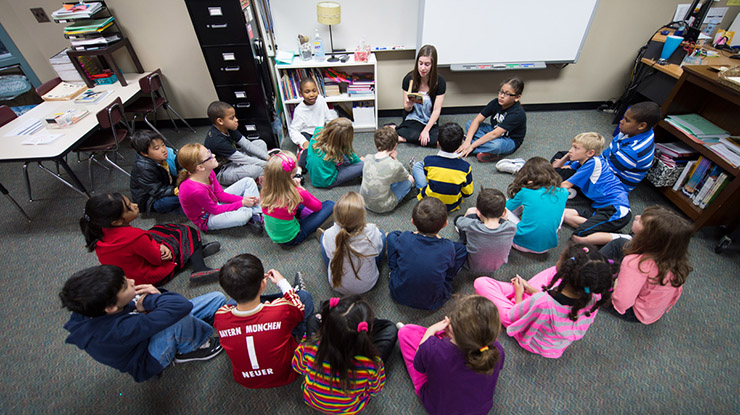 A student working on an elementary education degree sits on the floor and reads a story to a classroom of children.