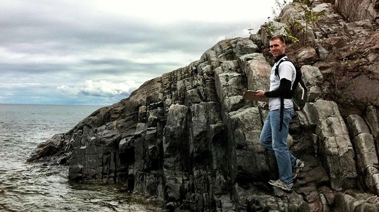 An MSU student pursuing a geology degree balances on the edge of rugged terrain next to an expanse of water and cloudy sky.