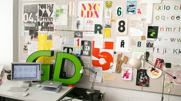A graphic design major’s workspace — a desk, computer and a variety of stylized letters and numbers on a bulletin board.