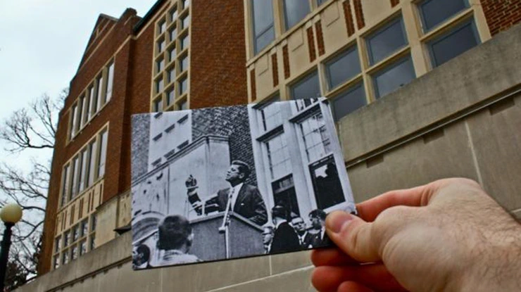 A history major holds a black-and-white photo of JFK speaking at a podium in front of a modern-day building.