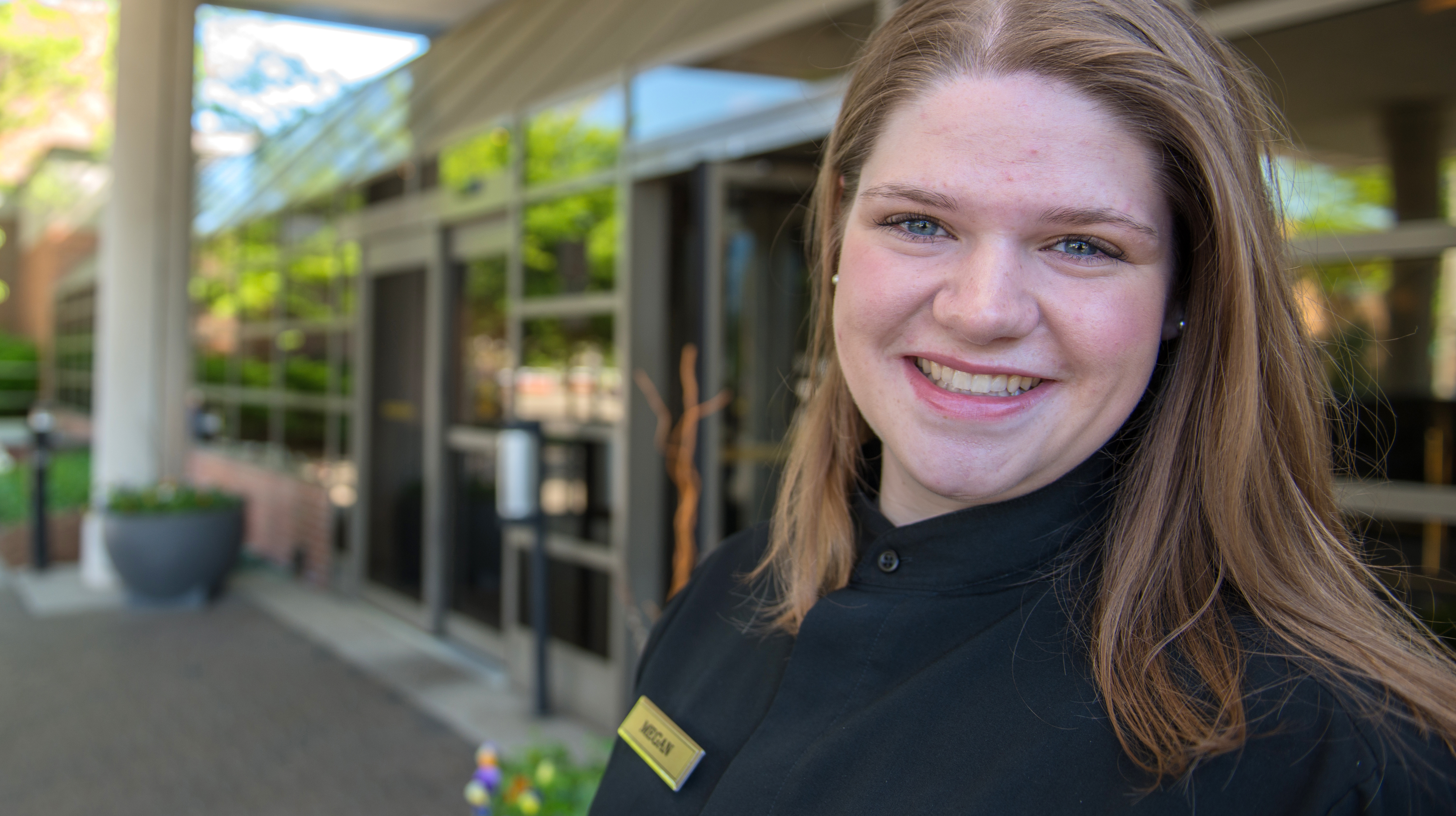 A smiling hospitality major stands outside the entrance to a hotel lobby, wearing black clothes and a name tag.