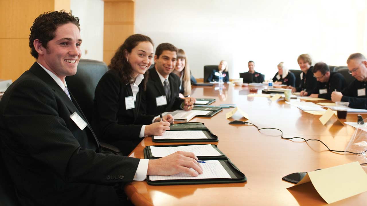 A group of students working on a marketing degree sit around a long table in a conference room wearing suits and nametags.