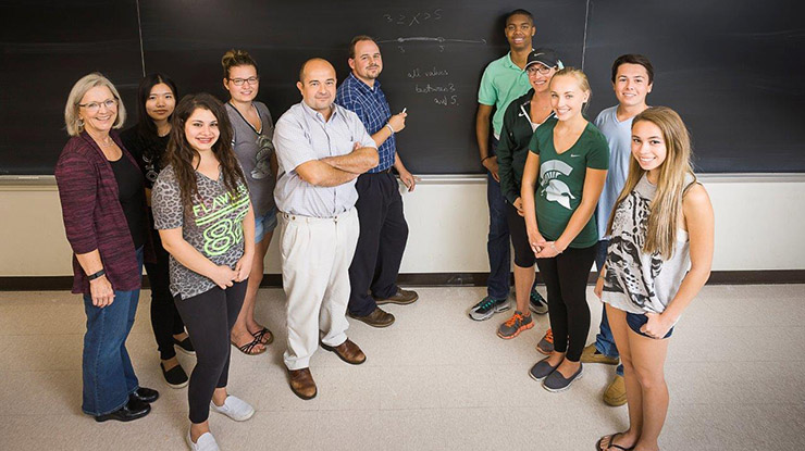 A group of smiling math majors and faculty members stand in front of a chalkboard.