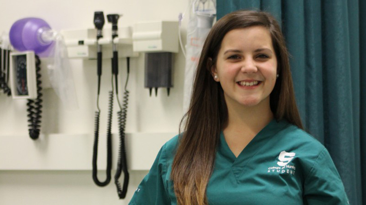 A nursing major wearing Michigan State scrubs smiles and stands in front of a wall of medical equipment.