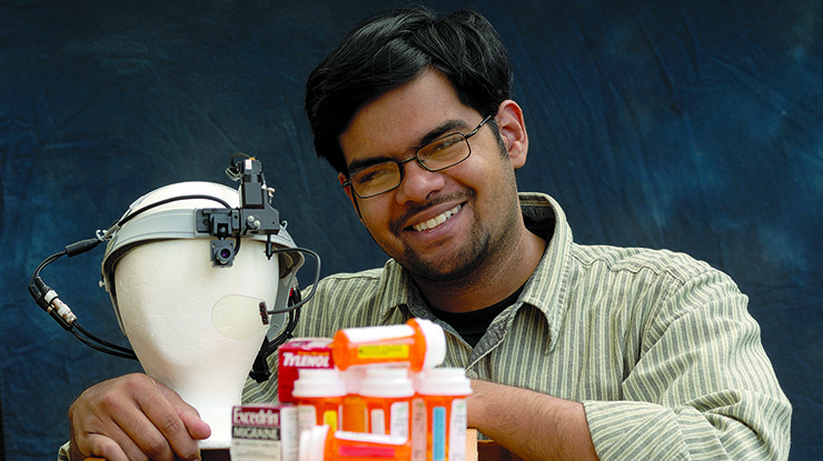 A smiling packaging major sits next to a mannequin head wearing a device, pill bottles and boxes of Tylenol and Excedrin.