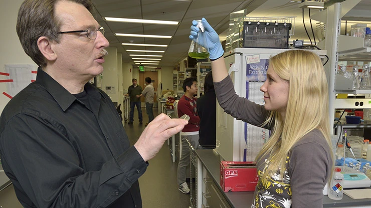 In a lab, a plant biology major holds up a beaker of green liquid while a professor explains a concept for class.
