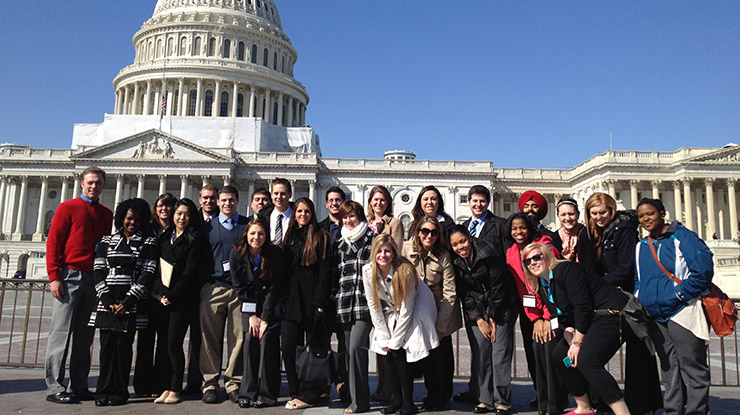 A large group of students working on a political theory degree smile as they stand in front of the US Capitol building.