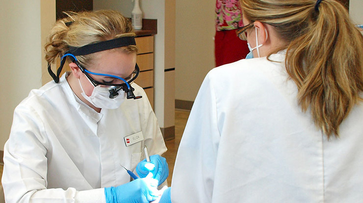 Two predental students wearing white lab coats and masks practice using dental equipment.