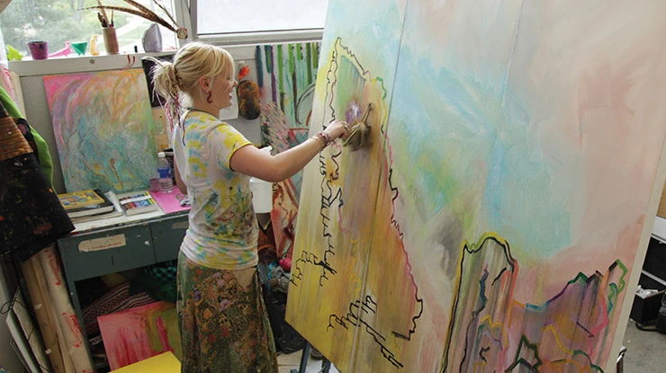 A studio art major in paint-splattered clothing paints on a large colorful canvas in an art studio.
