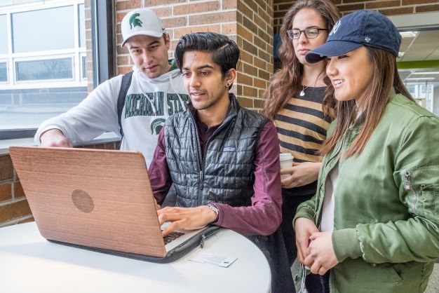 Students gathered around a laptop in the Communication Arts and Sciences Building at Michigan State University)