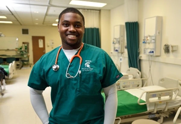 An MSU nursing student wearing green scrubs in a hospital setting smiling with a stethoscope draped over their neck 