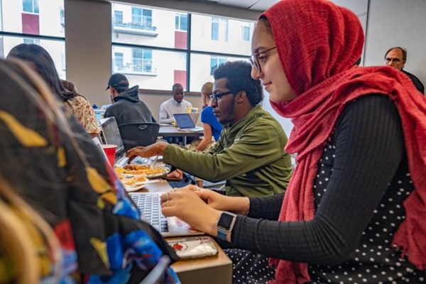 A student wearing a vibrant red hijab and polka dot outfit types on a laptop in a classroom full of students.