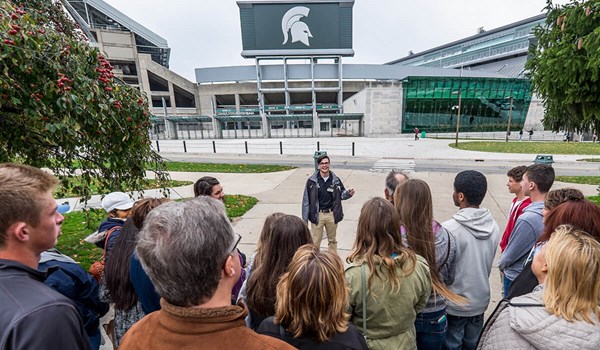 A student tour guide leads a tour group standing in front of Spartan Stadium at Michigan State University