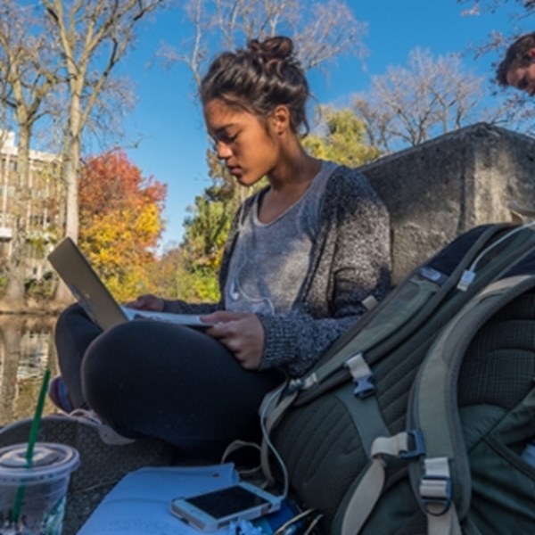 A student sits by the Red Cedar River and reads on her laptop