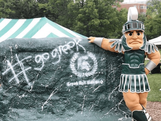 Michigan State University mascot, Sparty, leaning against the rock that is painting green with "# go greek" written in white